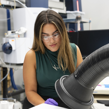 A woman with long dirty blonde hair, wearing a green blouse and safety glasses works in a lab with a large black hose.