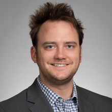 Professional headshot of Zachary Norris Buck, a young man with messy hair, smiling, wearing a collared shirt and suit jacket.