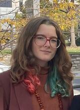 Headshot of Katarina Goodge, an individual with long brown hair with green and red tips, wearing glasses.