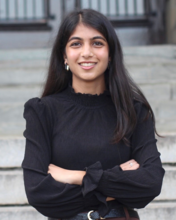 Portrait of Radha Patel, a young lady with long dark hair, wearing a black blouse, smiling for a photo in front of steps.