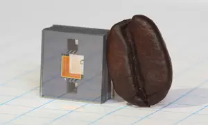 Square gray cube with two square holes in it and a gold square in between the holes, and a dark brown coffee bean leaning against the cube.