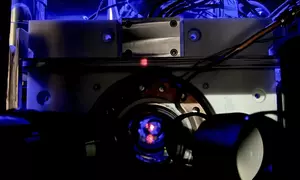 Close-up of a scientific instrument with wires and a circular metallic component, illuminated by blue light, featuring a small red dot at the center.