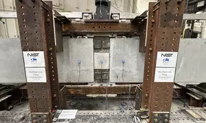 A wide view of a concrete beam assembly inside a large metal frame. A hydraulic arm is part of the metal frame, and it’s posed to press down at a section in the middle where the concrete pieces connect.