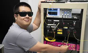 Yicheng Shi wears dark safety glasses as he poses next to a rack of electronics equipment.