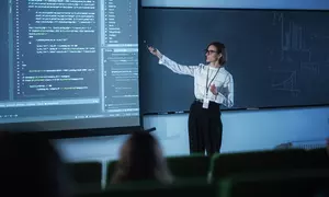 A female instructor wearing glasses presents code on a large screen to a classroom.