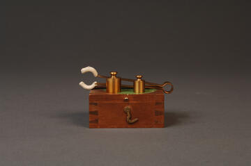 Two gold-plated brass weights sitting on top of a wood box, in front of tongs used to handle them