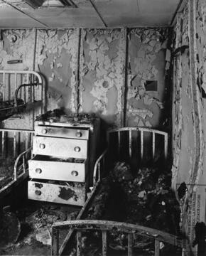 Black and white photo of post fire aluminum bedroom replica of SS united States ship in 1950. To the right and left there are burned bed frames and in the middle a burned dresser.