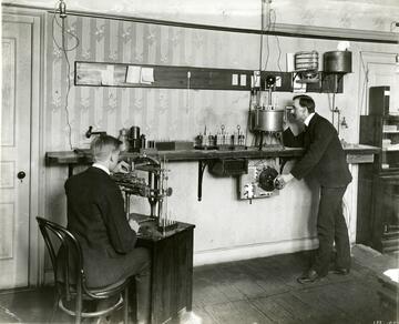 Man sits at a desk on the left while a man on the right stands looking into a device