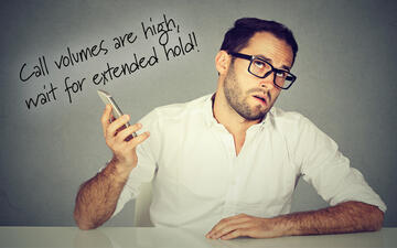 Frustrated man holding a cell phone listening to automated message saying "Call volumes are high, wait for extended hold!"
