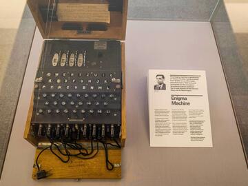 Left: Machine that looks like an old fashioned typewriter in a box. Right: picture of Alan Turing along with a bio. Both are from a museum exhibit.