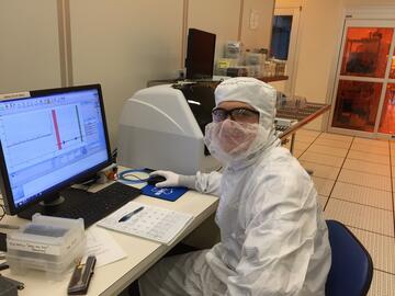 A person in a white coverall and safety glasses poses sitting at a computer in a lab.
