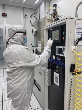 A person in a white coverall and mask stands at a screen on a large piece of equipment in a lab. 