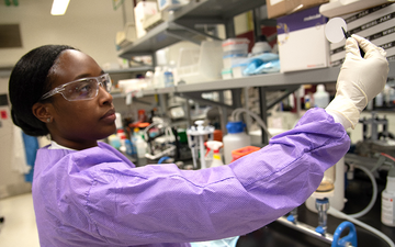 Alshae' Logan-Jackson wears a purple coverall and safety glasses in the lab as she uses tweezers to hold a small white disk. 