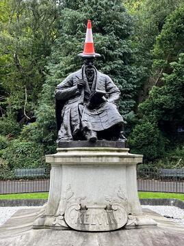 A statue of Lord Kelvin, seated with a notebook, stands in a leafy park. An orange traffic cone is placed on his head like a hat.  