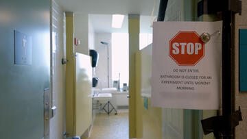 A paper sign is clipped in front of a open door to a public restroom. The sign reads, “Stop. Do Not Enter. Bathroom is closed for an experiment until Monday Morning.”