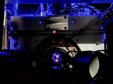 Close-up of a scientific instrument with wires and a circular metallic component, illuminated by blue light, featuring a small red dot at the center.