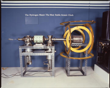 Museum display shows a cylindrical device lying horizontally on two mounts. Text on wall reads: The Hydrogen Maser: The Most Stable Atomic Clock. 