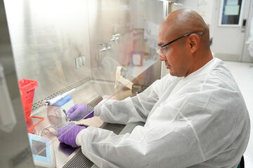 A researcher wearing safety gear works with small tools in the lab, putting his hands inside a plexiglass box. 