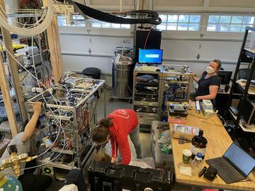 The inside of a two-car garage is filled with racks of equipment and wiring, with three researchers working to arrange the equipment. 
