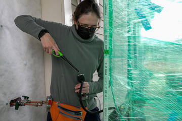 A researcher standing outside a house holds a lighter to the end of a hose-like device leading into an area closed off by plastic wrap. 