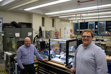 Three men in safety glasses pose standing around a large clear plastic box on a lab table. The box holds several small pieces of equipment. 