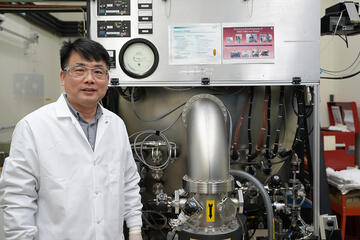 A researcher in a lab coat and safety glasses poses next to a large piece of lab equipment.