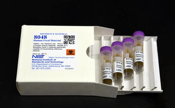 A white box labeled "8048 Human Fecal Material" lies open, with a tray of four plastic vials pulled out. 