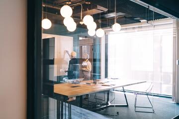 At the far end of a modern conference room with overhead globe lighting, two people stand looking at a whiteboard. 