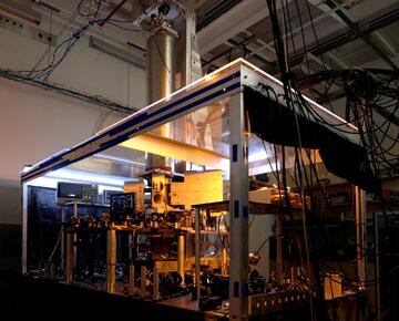 A large clear-sided box inside a darkened lab holds electronic equipment, wiring, and a metal cylinder rising up from the box. 