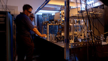 Two researchers in safety glasses stand at the edge of a metal table holding electronic and laser devices, wiring and metal structures.