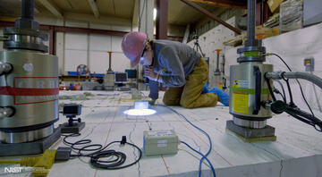 A researcher in a hard hat kneels on a concrete slab and uses a flashlight to examine it closely.