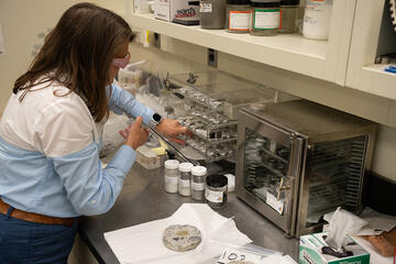 A researcher wearing safety glasses leans over the lab table near where a small disk of concrete is sitting. 