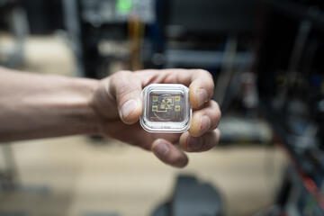 A hand holds a small square device in a clear case. Gold-colored electronics can be seen through the case. 