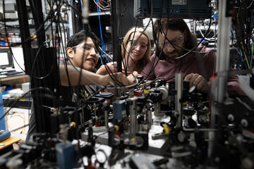 Three researchers in safety glasses are seen through a forest of hanging wires and other electronic and laser devices. 