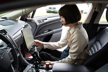 A woman sits in the passenger side of a parked car, looking at a large monitor attached to the dashboard. 