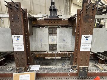 A wide view of a concrete beam assembly inside a large metal frame. A hydraulic arm is part of the metal frame, and it’s posed to press down at a section in the middle where the concrete pieces connect.