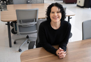 Kelly Sauerwein poses sitting at a table in the NIST library, leaning forward and smiling. 