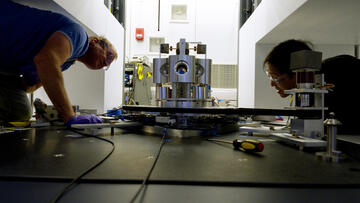 Wide shot shows two researchers on either side of a scientific device made of metal cylinders, peering closely at it. 