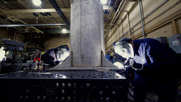 Two people wearing welding masks and other safety equipment bend over a table to work on a large metal cylinder.