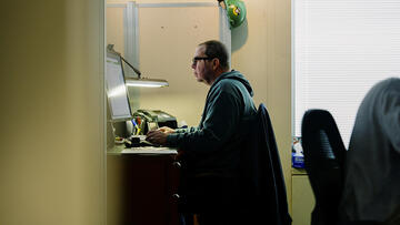 Side view shows a man sitting at a desk, working on a computer in a small, shared office.