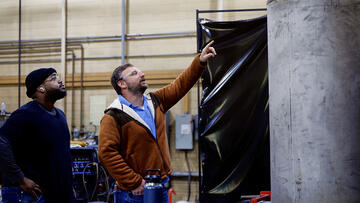 Two men in safety glasses stand in the lab looking at a large metal cylinder.