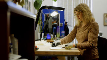 A woman wearing special gloves stands at a high table, sorting small pieces of paper. 
