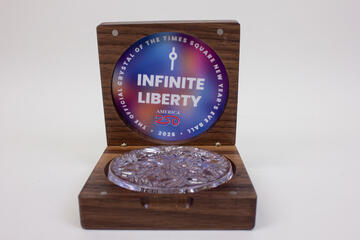 A clear crystal sits in a wooden box. On the top center of the box is a label that reads  "The Official Crystal of The Times Square New Year's Eve Ball 2026"