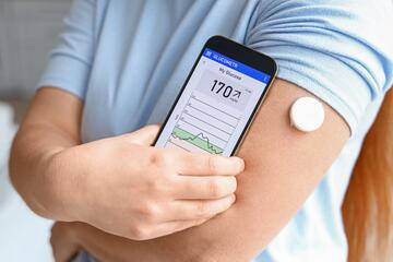 Close-up view of a woman holding a phone up to the round white device attached to her arm. The phone shows chart labeled "My Glucose." 