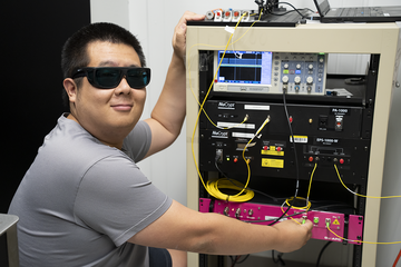 Yicheng Shi wears dark safety glasses as he poses next to a rack of electronics equipment.