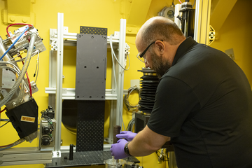 A man in safety glasses carefully handles a dark scroll on a table surrounded by imaging equipment. 