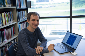 Cyrus Daugherty sits smiling at a table in a sunny library, with a laptop showing a spiral image.