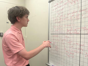 Henry Nonnemaker stands holding a marker, looking at a gridded whiteboard covered with mathematical formulas.