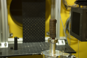 A charred, rolled-up scroll stands vertically on a metal holder surrounded by imaging equipment. 