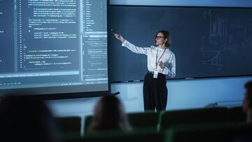 A female instructor wearing glasses presents code on a large screen to a classroom.